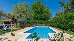 Une piscine rectangulaire bordée de carreaux, entourée d’une terrasse en béton. On y trouve des chaises longues et un espace barbecue doté d’un toit carrelé.