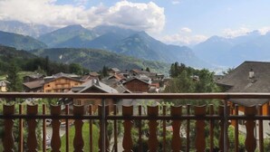 Un village de montagne aux maisons traditionnelles en bois, doté d’un balcon orné d’une balustrade en fer forgé, et offrant une vue sur les montagnes lointaines sous un ciel partiellement nuageux.