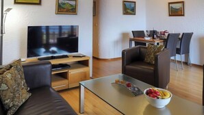 Un salon moderne avec un téléviseur à écran plat, un canapé en cuir et une table basse en verre sur laquelle sont disposés des fruits.