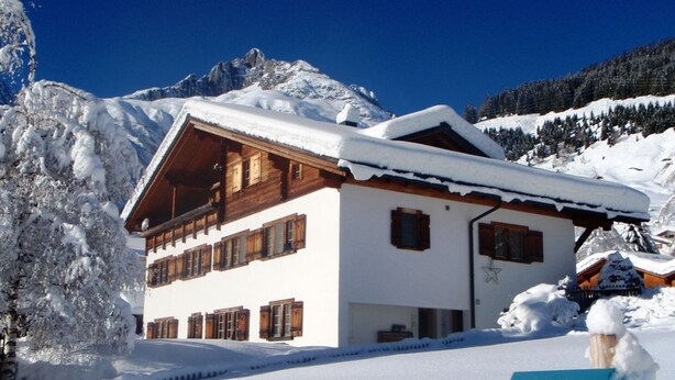 Ein schneebedecktes Chalet mit Holzfassade und zahlreichen Fenstern, umgeben von schneebedeckten Bäumen und Bergen.