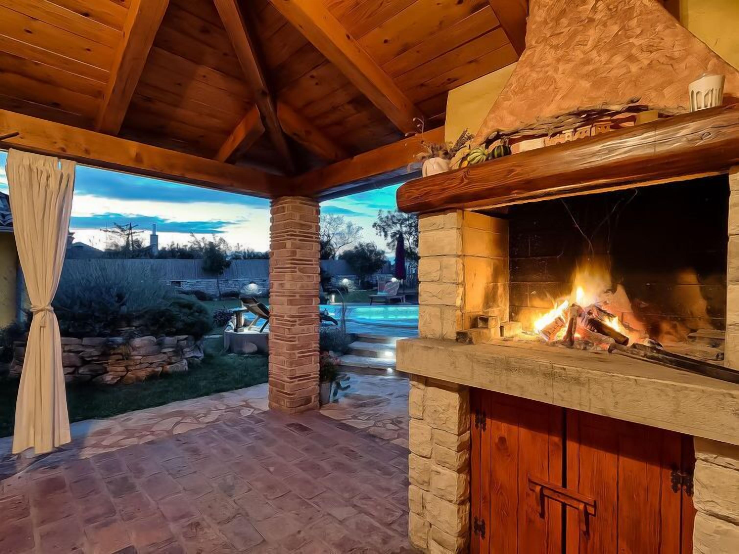 Ein Steinkamin mit Feuer, eine überdachte Terrasse und ein Pool im Hintergrund.