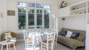 A bright room with a large window, a dining table with chairs, a sofa, and a small side table with a plant. - Apartment Du lac by Interhome (Hossegor)
