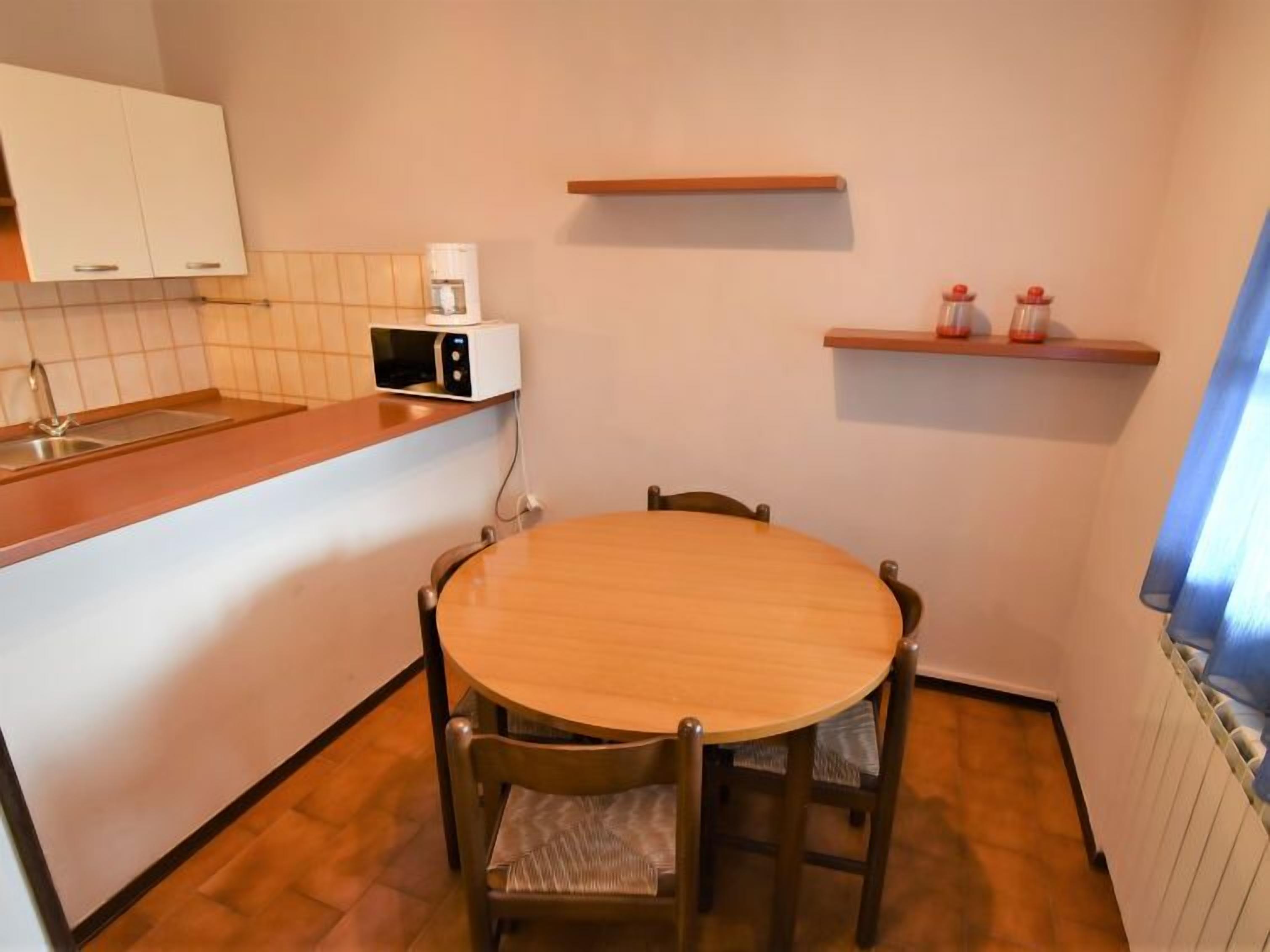A compact kitchen with a round wooden table, four chairs, a microwave, a shelf with bottles, and a window with curtains.