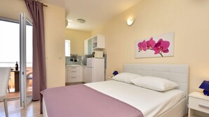 A hotel room with a bed, a nightstand, a wall-mounted lamp, a painting of pink flowers, and a view of the kitchen and balcony. - Melandura by Interhome (Pag/Dinjiška)