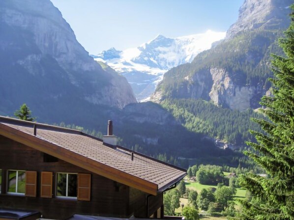 A mountain cabin with a sloped roof and a chimney, overlooking a valley with dense forests and snow-capped mountains.