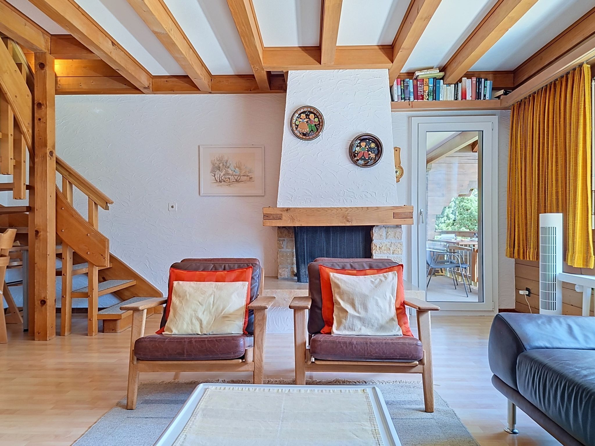 Ein Wohnzimmer mit Kamin, Holzbalken, einer Treppe und Blick ins Freie.