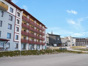 Un edificio residencial de varios pisos con balcones, un estacionamiento y un complejo de esquí al fondo.