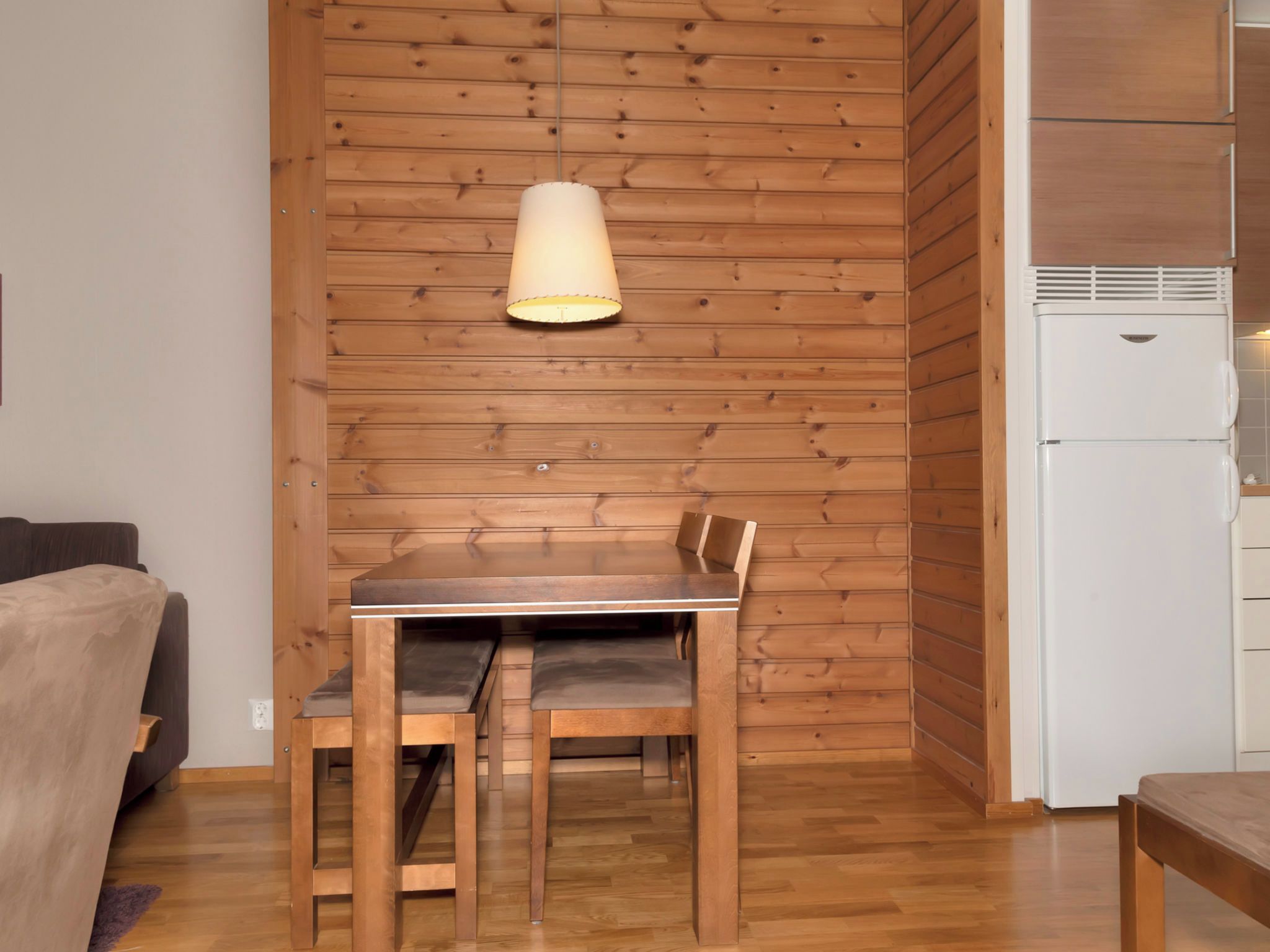 A dining area with a wooden table and chairs, a hanging light fixture, and a white refrigerator.