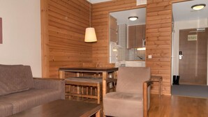 A cozy living room with a wooden wall, a sofa, a coffee table, and a chair. A kitchen is visible through an open doorway.