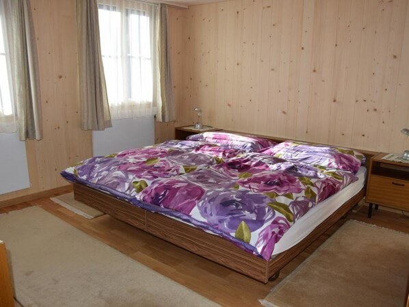 A bedroom with a wooden bed, a nightstand, a lamp, and a window with curtains. - Däpp # 2 by Interhome (Lenk)