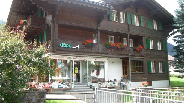 A traditional wooden building with a shop on the ground floor, balconies with flowers, and a stone wall in the foreground. - Däpp # 2 by Interhome (Lenk)