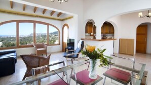 Uma sala de estar espaçosa com uma grande janela que oferece uma vista panorâmica, uma área de jantar com mesas de vidro e uma cozinha com armários de madeira.