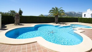 Uma piscina com formato singular, rodeada por um deck revestido de azulejos e palmeiras.