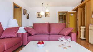 Un salon avec un canapé rouge, une table basse blanche sur laquelle se trouve un saladier rempli de fruits et des verres à vin, ainsi qu’une lampe.