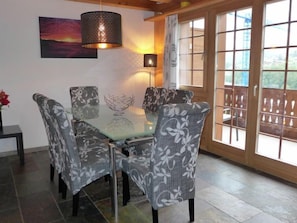 A dining area with a glass table and patterned chairs, a pendant light, and a view through large windows.