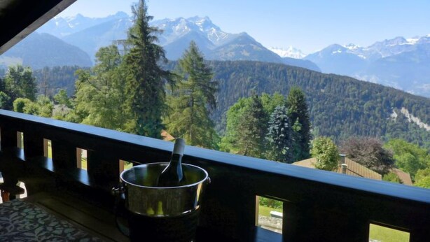 A balcony with a metal bucket and a spoon, overlooking a mountainous landscape with trees and snow-capped peaks. - Regina B9 by Interhome (Villars)