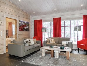 Una sala de estar moderna con dos sofás, una mesa de centro y una silla roja. Hay ventanas grandes con cortinas rojas, un reloj en la pared y un cuadro sobre el sofá.