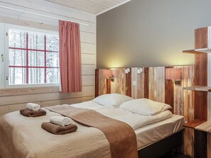 Un dormitorio con cabecera de madera, cama con ropa de cama blanca y ventana con vistas a árboles cubiertos de nieve.
