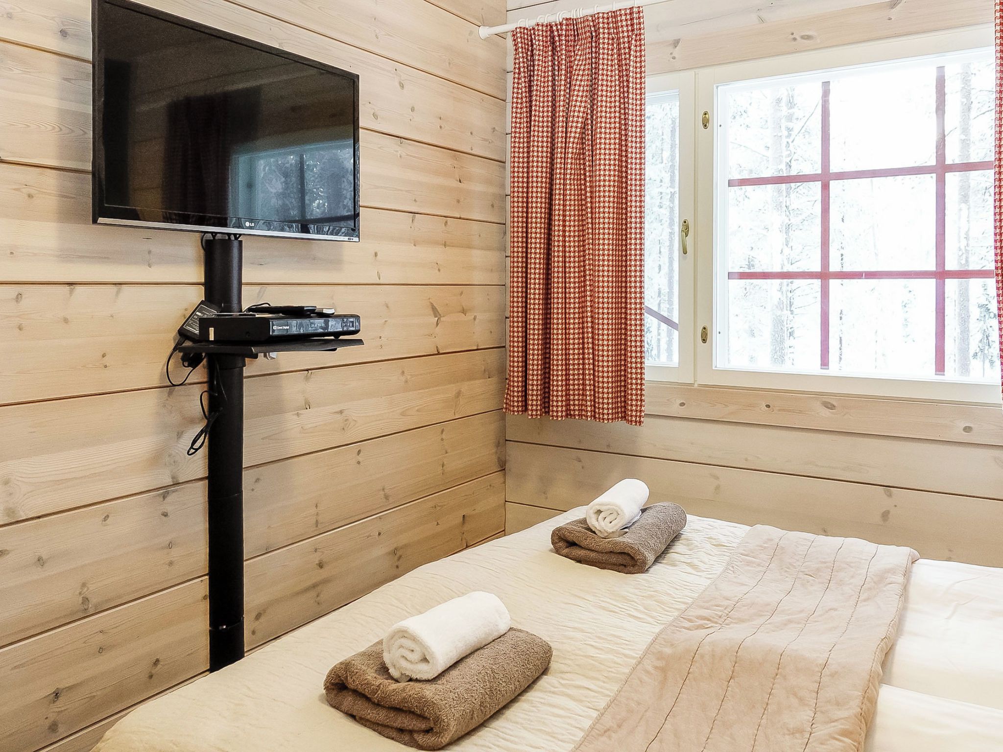 Een kamer met houten wanden, een flatscreen televisie op een standaard, een raam met een rood-wit geruit gordijn en twee bedden met handdoeken.