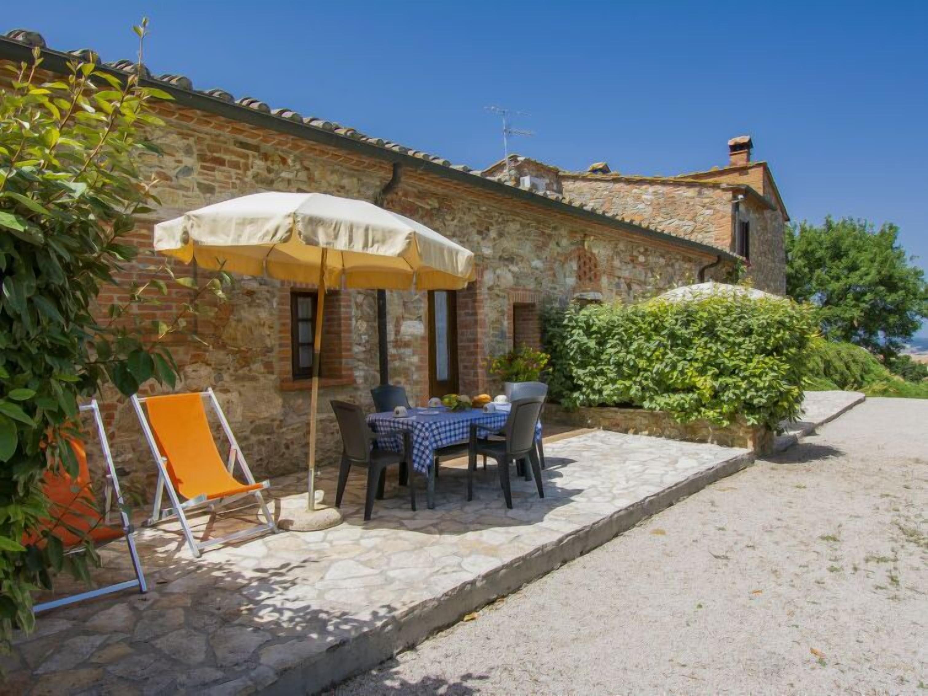 Un bâtiment en pierre avec une table et des chaises placées sous un parasol jaune, un petit jardin et un ciel bleu dégagé.