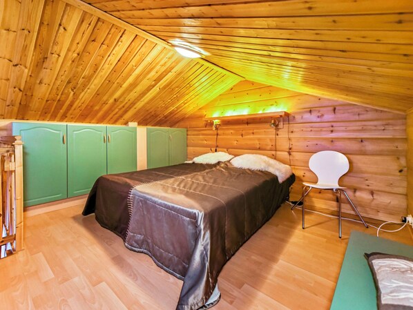 Un dormitorio acogedor en el ático con techo de madera, una cama con colcha oscura, un armario verde y una silla blanca.