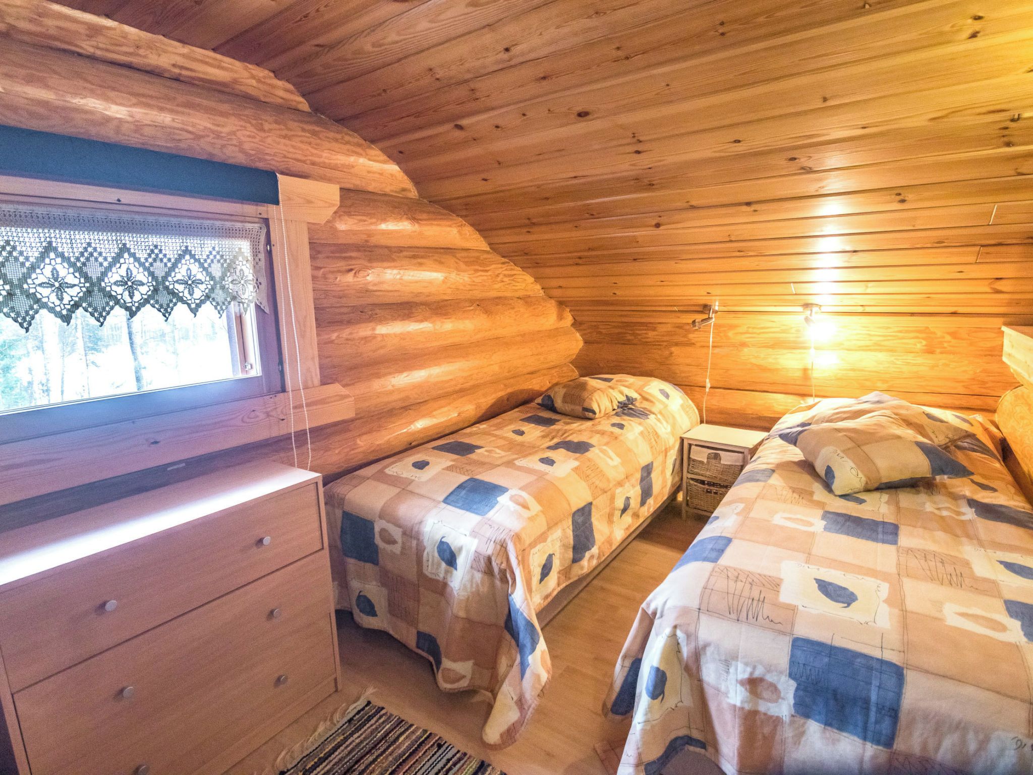 A cabin with a wooden interior, a window with lace curtains, two beds with checkered bedding, and a wooden dresser.