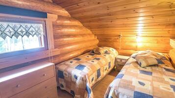 A cabin with a wooden interior, a window with lace curtains, two beds with checkered bedding, and a wooden dresser.