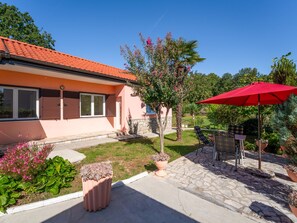 Une maison avec un toit recouvert de tuiles rouges, un jardin avec différentes espèces de plantes, une terrasse avec des meubles d’extérieur et un parasol rouge.
