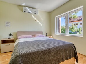 A bedroom with a bed, bedside tables, an air conditioning unit, and a window with a view of buildings and trees. - Vacation home Franjo by Interhome (Opatija/Ika)