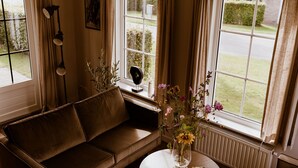 Ein gemütliches Wohnzimmer mit einer braunen Ledersofa, einem runden Couchtisch mit Blumen und Blick auf einen grünen Garten durch große Fenster.