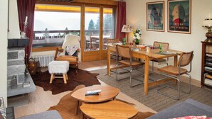 A cozy living room with a fireplace, a dining area with a wooden table and chairs, a sofa, and a view of the outdoors through large windows. - Schuss 23 by Interhome (Villars)