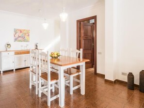 Un comedor con una mesa de madera y sillas blancas, un armario blanco con un florero, y un cuadro en la pared.