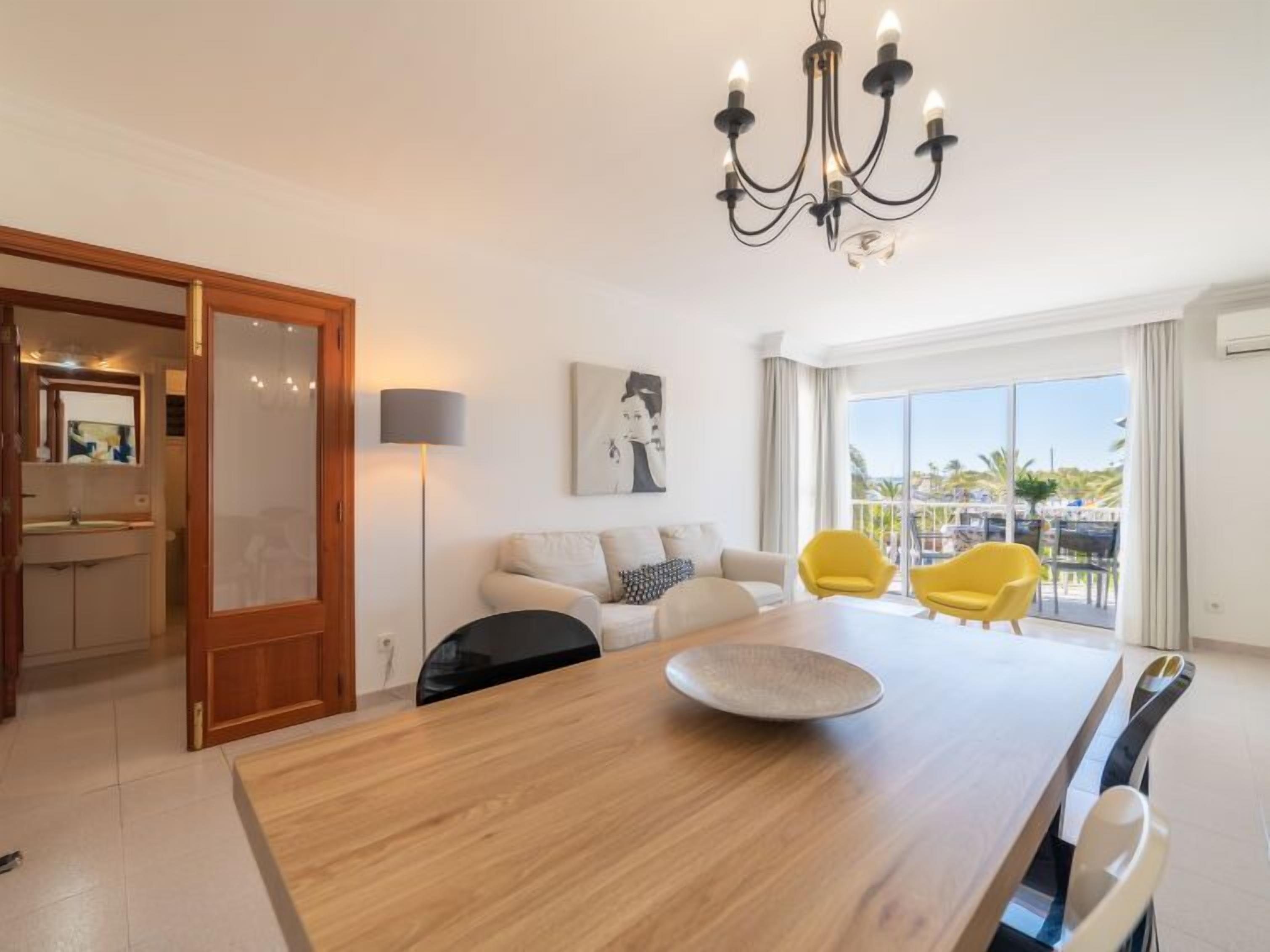 Un salon moderne avec une table à manger en bois, un canapé et deux fauteuils jaunes. Un lustre est suspendu au plafond, et une grande fenêtre offre une vue sur les palmiers.
