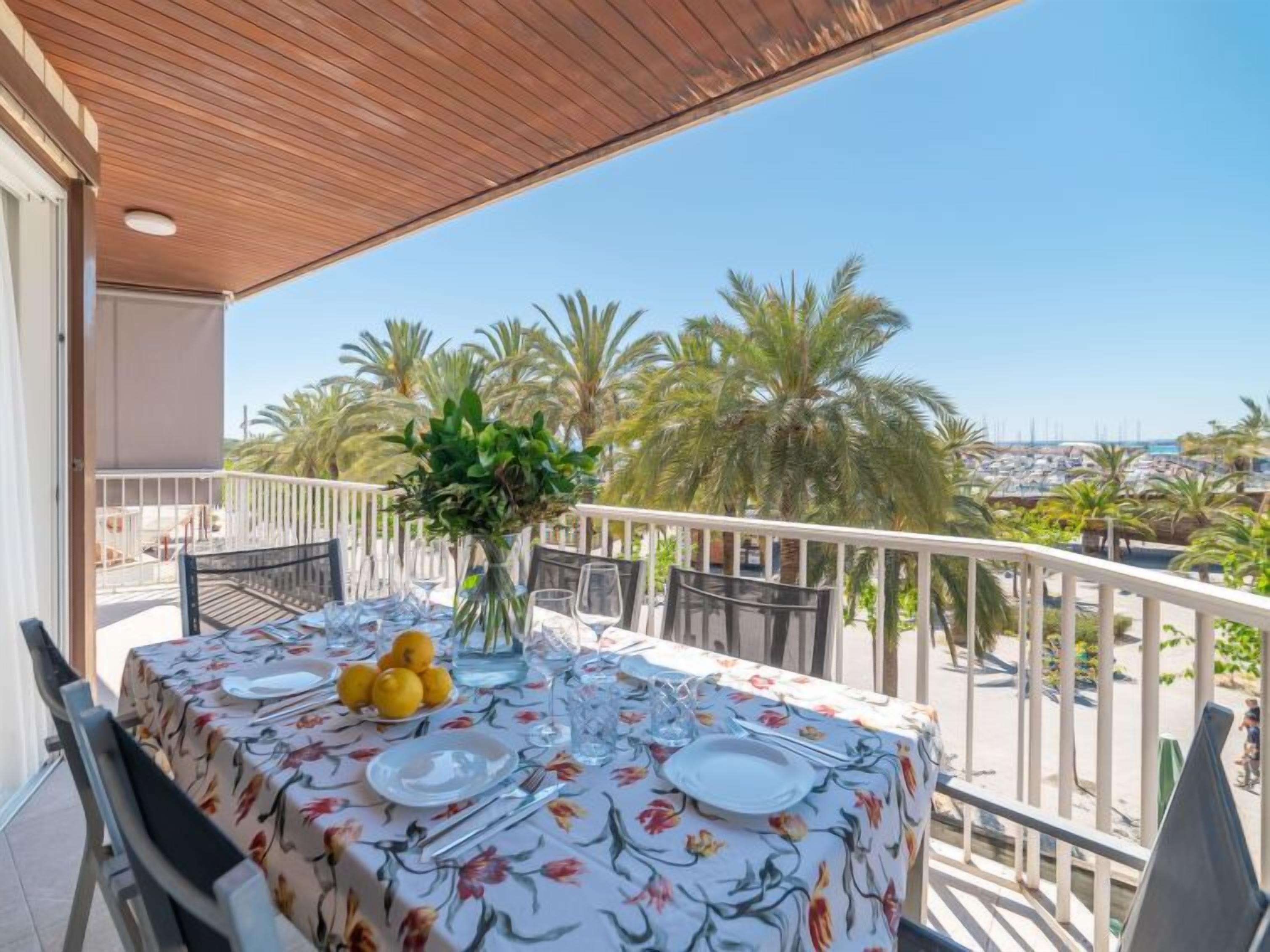 A table set for two with a floral tablecloth, a vase with greenery, and a bowl of lemons on a balcony with a view of palm trees and a marina.