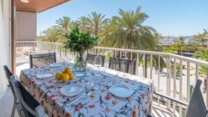 Une table dressée pour deux personnes, recouverte d’une nappe à motifs floraux, agrémentée d’un vase avec des plantes vertes et d’un bol de citrons, sur un balcon offrant une vue sur des palmiers et une marina.