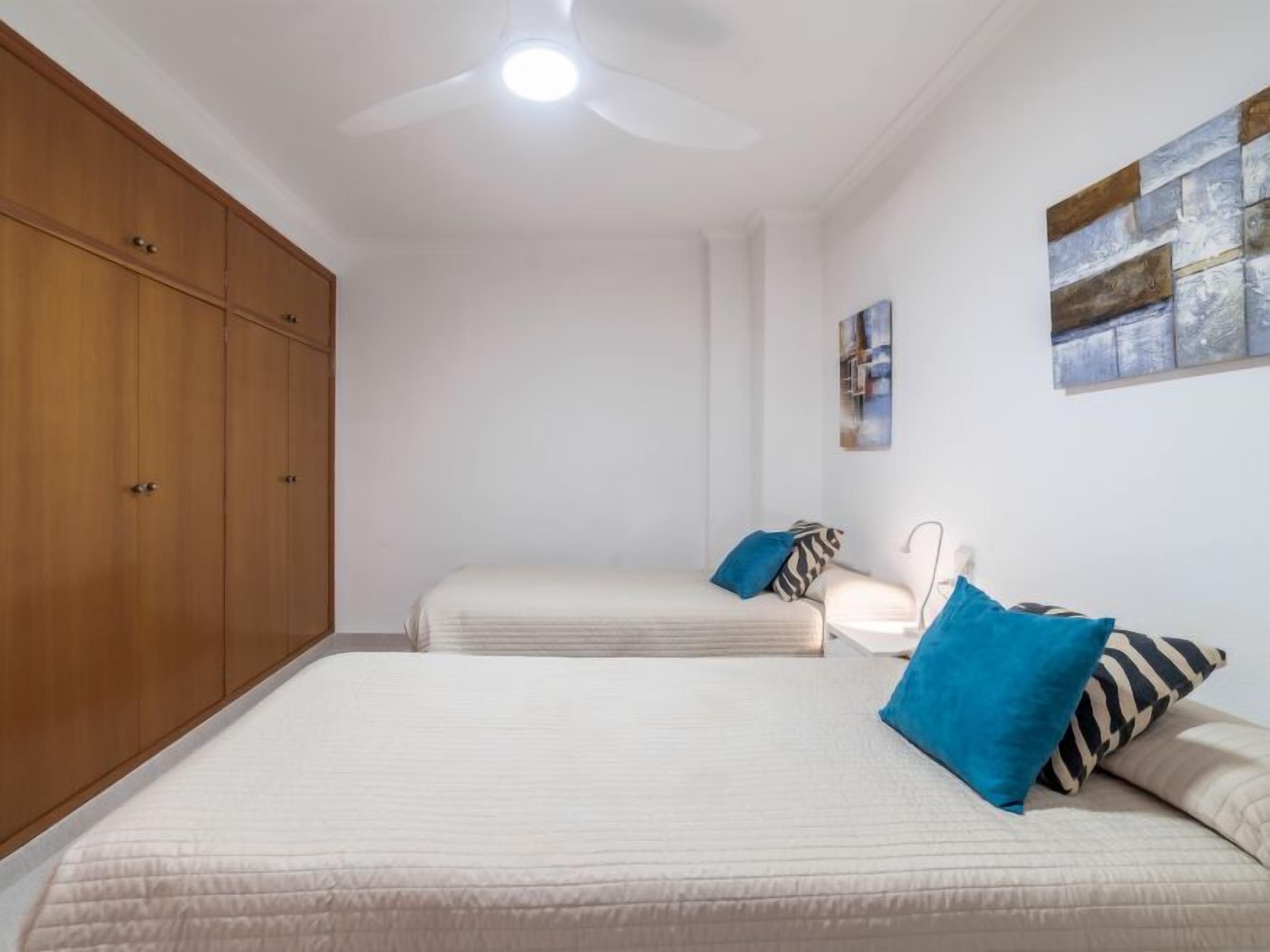 A bedroom with two beds, wooden wardrobe, ceiling fan, and artwork on the wall.