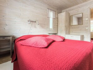 Een slaapkamer met een houten plafond, een bed met rode beddengoed, een nachtkastje en een raam met gordijnen.