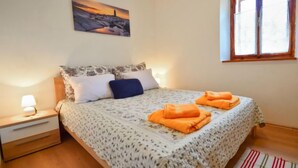 Un dormitorio con cama, mesita de noche, lámpara, ventana y un cuadro en la pared.