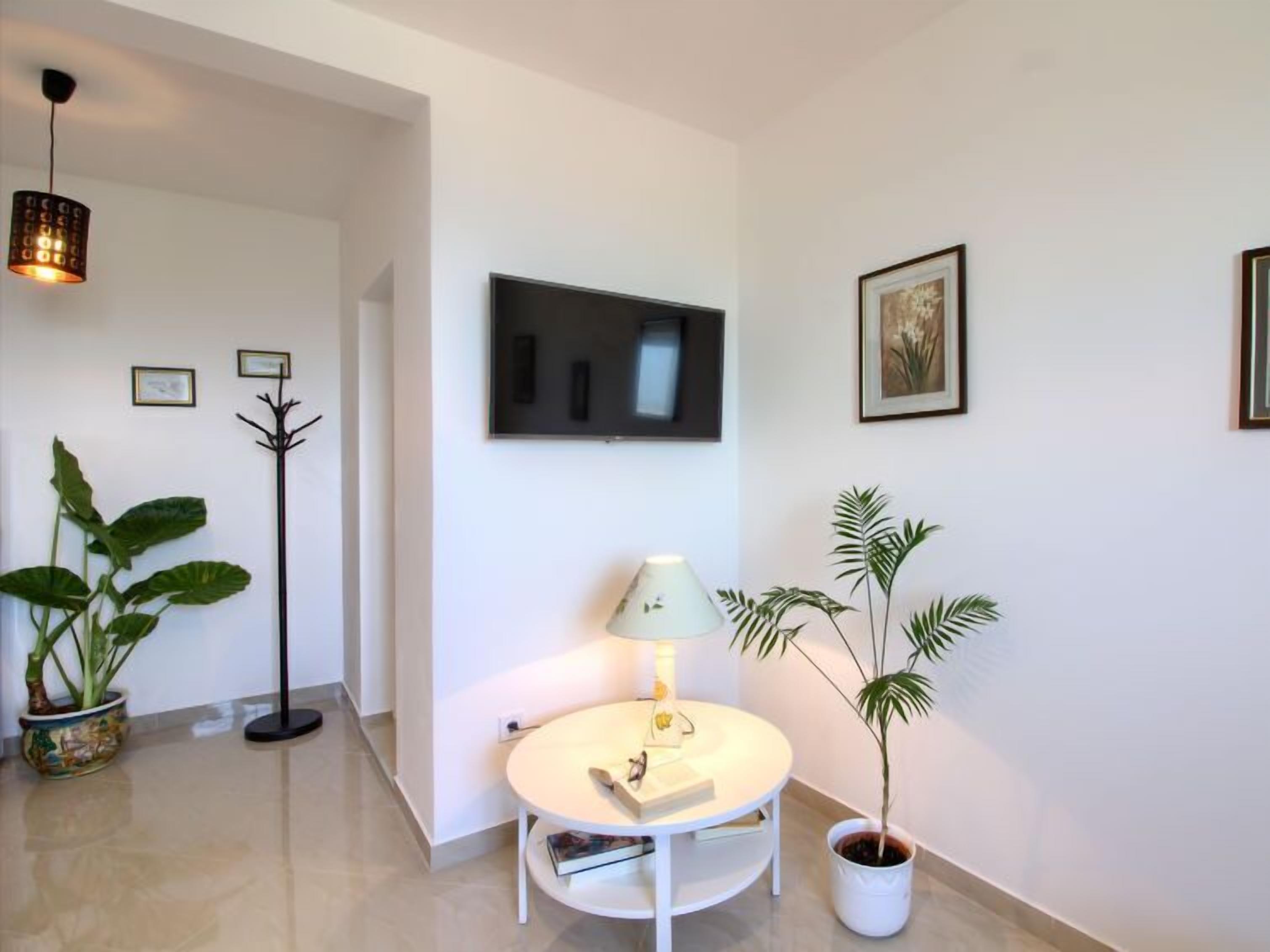 A modern living room with a flat-screen TV mounted on the wall, a round white table with a lamp, and two potted plants.