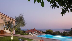 Zona junto a la piscina con sillones de descanso, una mesa con bebidas y vista al mar.