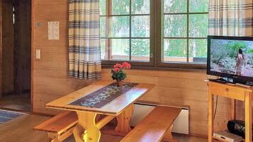 Un interior de madera con una mesa, bancos, un televisor sobre un soporte y una ventana con cortinas.