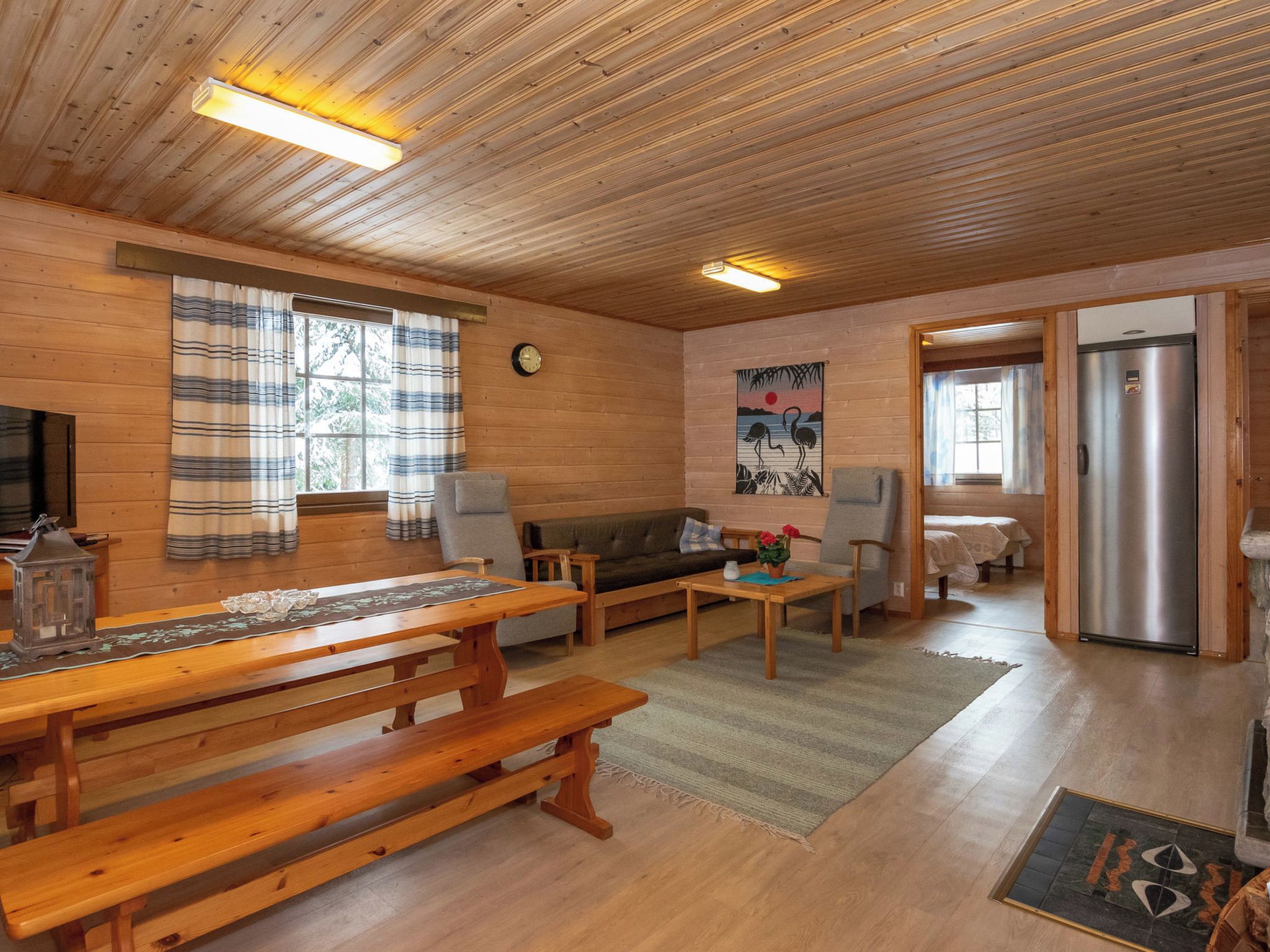 A cozy cabin interior with a wooden ceiling, a sofa, a dining table, and a refrigerator.
