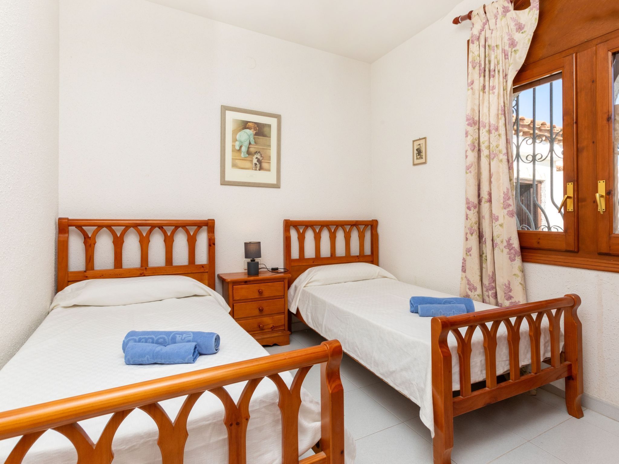 Un dormitorio con dos camas de madera, una mesita de noche, una lámpara y un ventanal con cortinas.