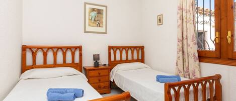 Un dormitorio con dos camas de madera, una mesita de noche, una lámpara y un ventanal con cortinas.