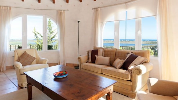 Een ruime woonkamer met een houten plafond, grote ramen met uitzicht op zee, een beige bank, een houten bijzettafeltje en een witte fauteuil.