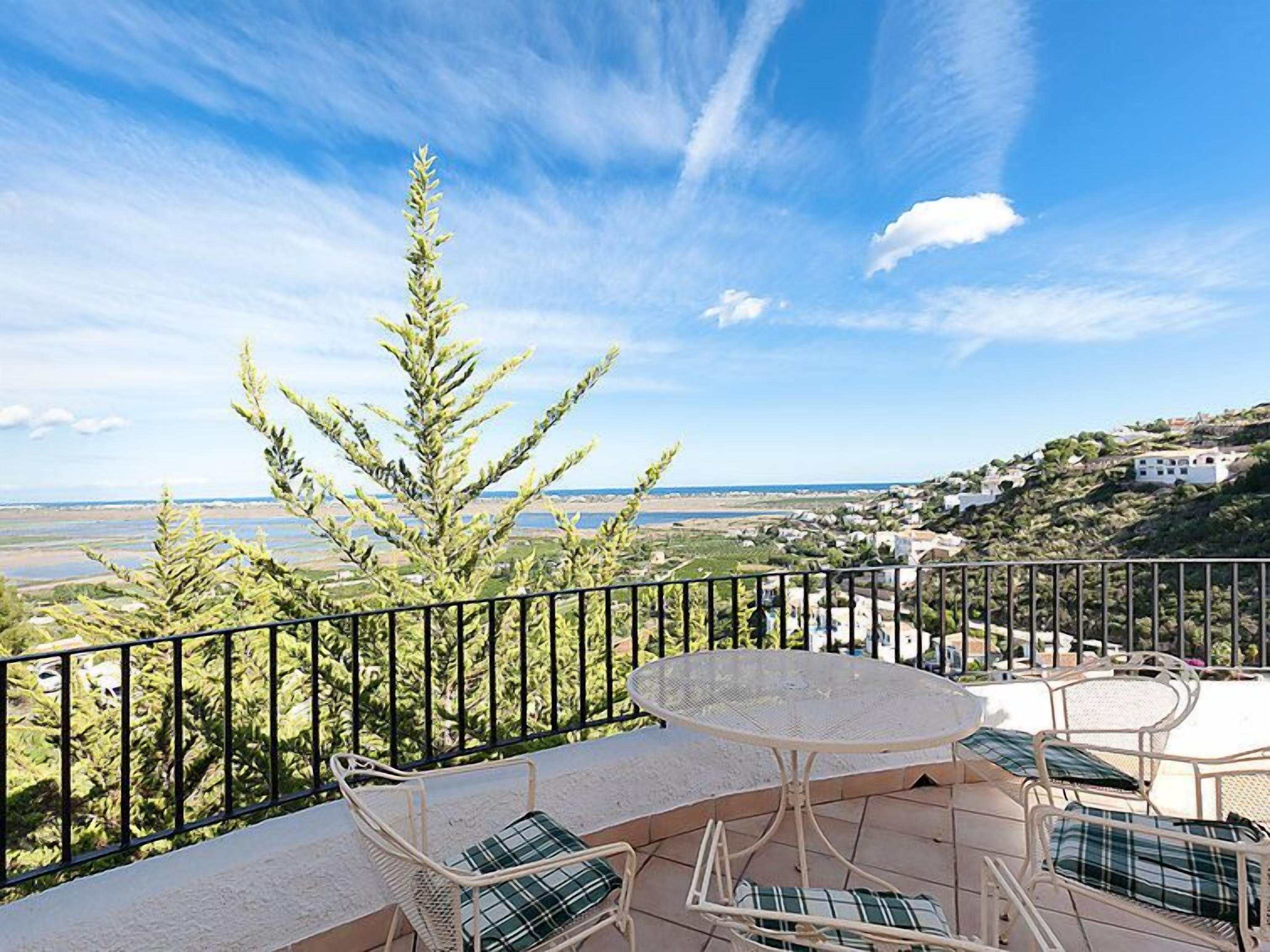 A balcony with a round table and chairs, overlooking a coastal landscape with a view of the sea and distant buildings.