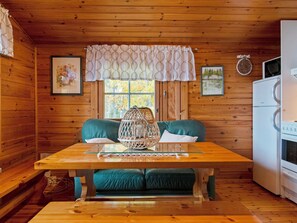 Interno di una baita in legno con un tavolo da pranzo, cuscini color acquamarina, un cesto intrecciato, opere d'arte incorniciate e un frigorifero bianco.