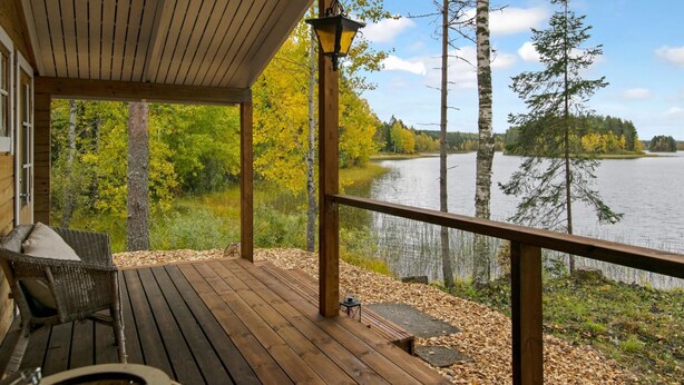 A wooden deck with a view of a lake and trees, a wicker chair, and a lantern. - Lahdenpohja, vaikon loma 7 by Interhome (Kaavi)