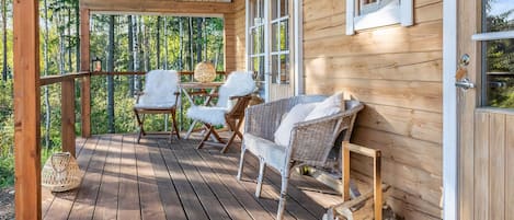 Una terraza de madera con techo, dos sillas blancas, un sofá de mimbre y una puerta con ventanal.