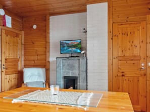 Interior de madera con televisor sobre una repisa, chimenea y mesa de comedor con un mantel individual.